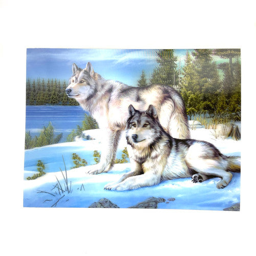 Two wolves relaxing on snow covered rocks by a frozen lake and pine trees.